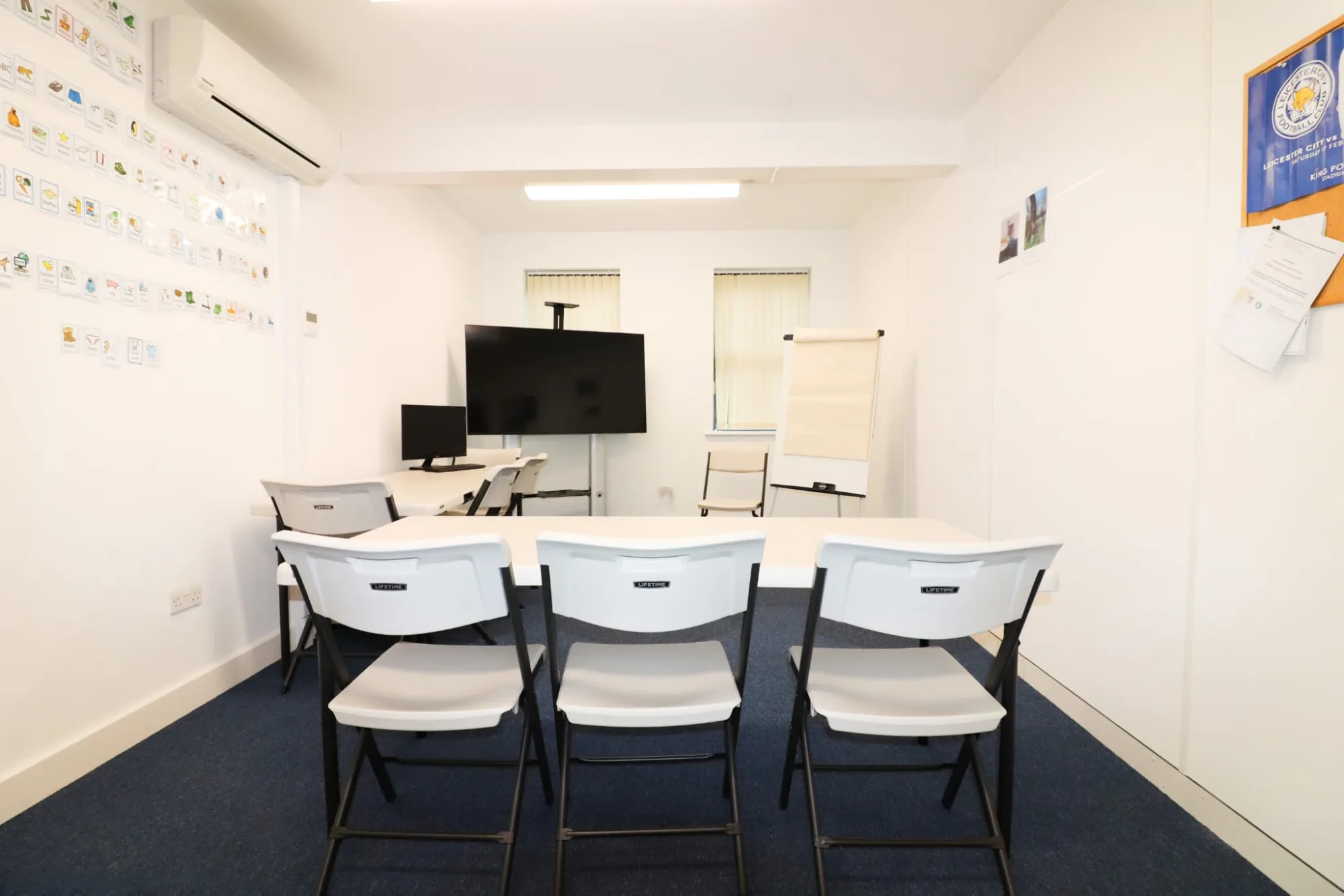 A small room with folding tables and chairs facing a screen and a flip chart, and a long wall of picture symbol cards to one side.