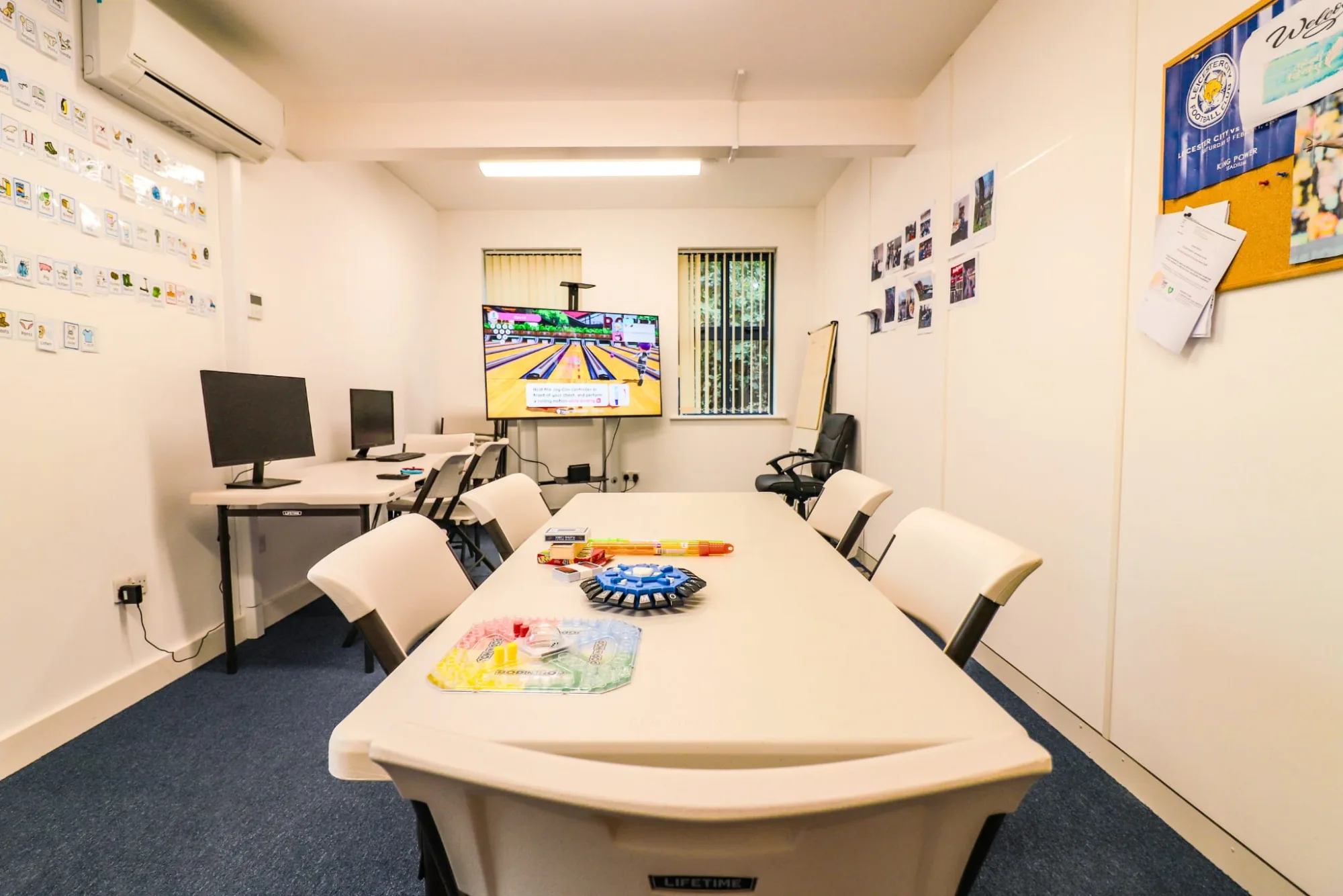 A long table set out with craft materials, two computers along the wall beside it and a screen at the far end showing a bowling game.