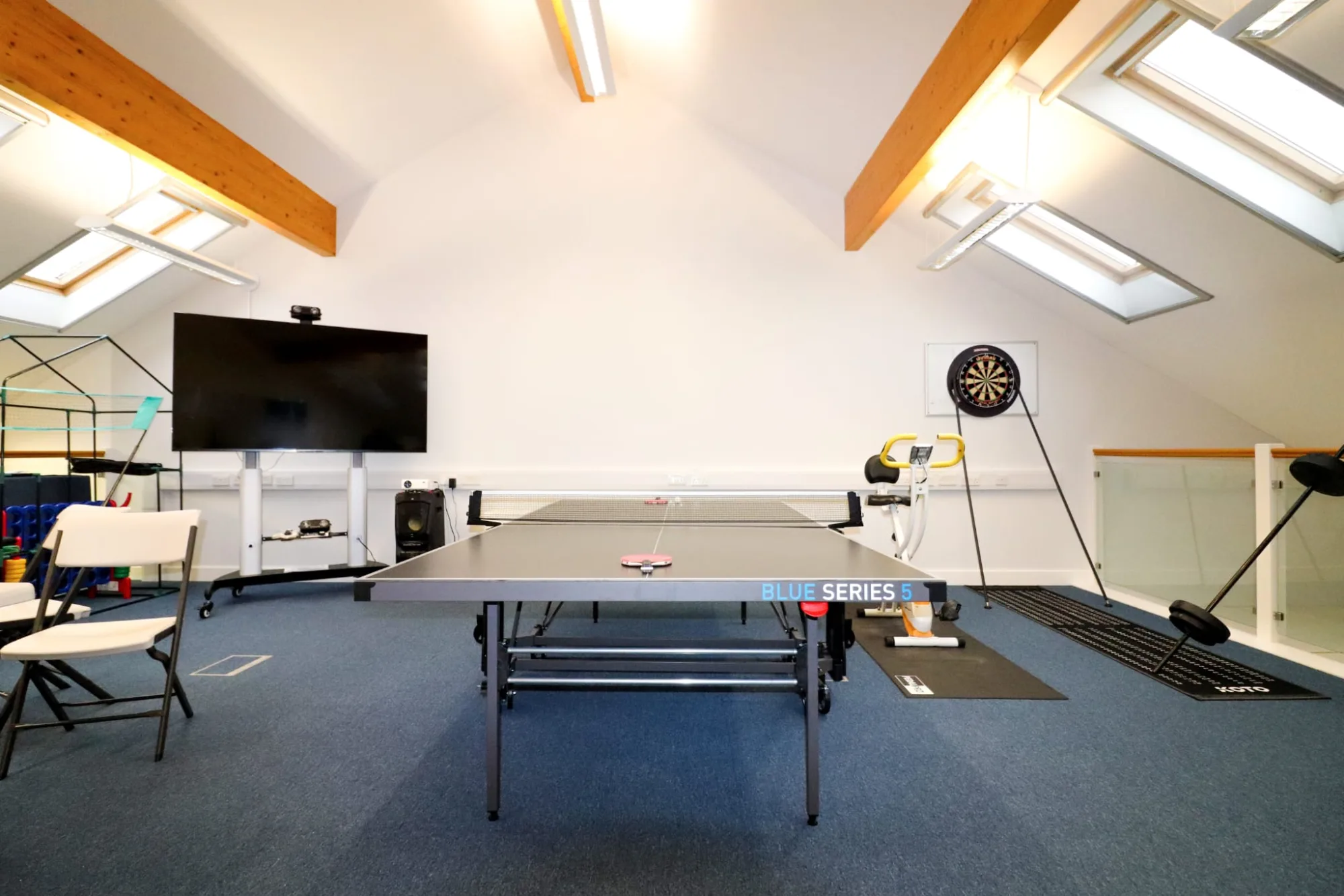 A large upstairs room under a pitched roof with skylights, a table tennis table in the middle, a wall-mounted screen on one wall and a dartboard on the other.
