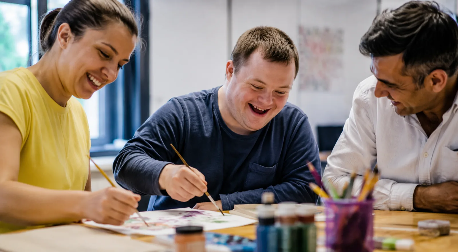 Three adults painting together at a table, laughing, with paints and brushes in front of them.